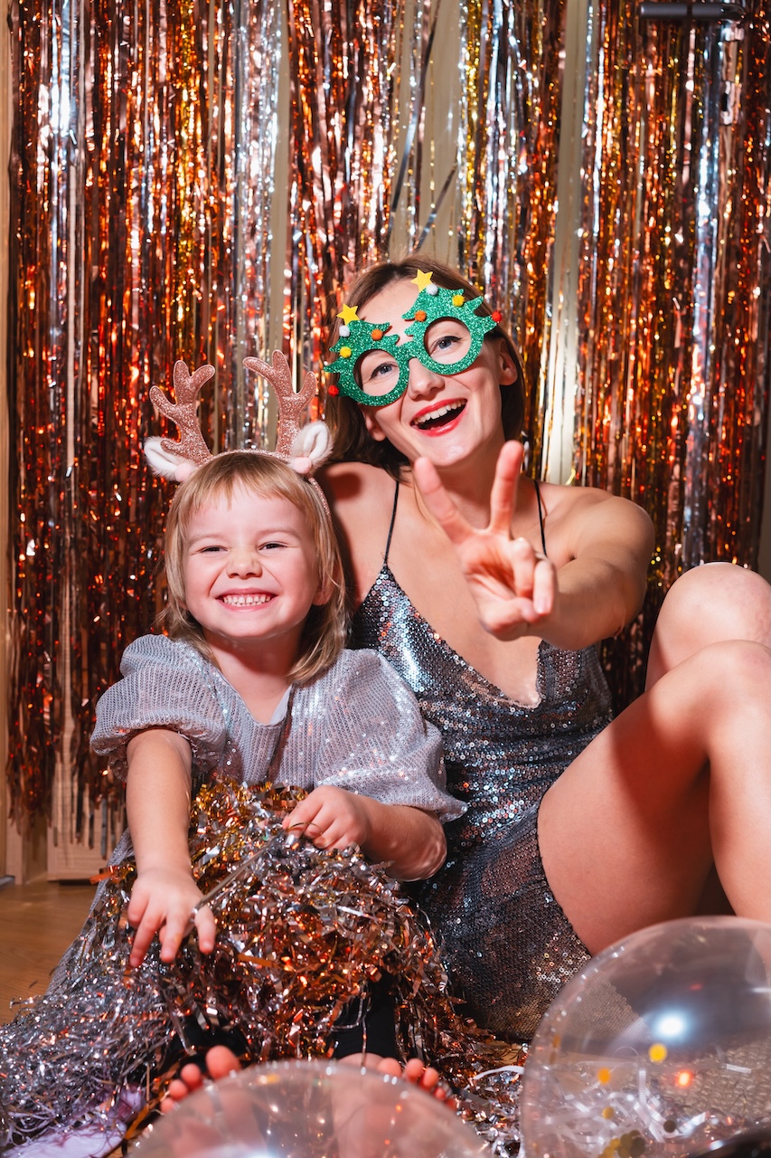 festive-girl-and-woman-celebrate-holiday-at-home-2026-03-19-10-37-15-utc Une femme vêtue d'une robe argentée et portant des lunettes de fête fait le signe de la paix à côté d'un enfant souriant coiffé de bois de renne ; tous deux sont entourés de guirlandes et de ballons, sur un fond scintillant.